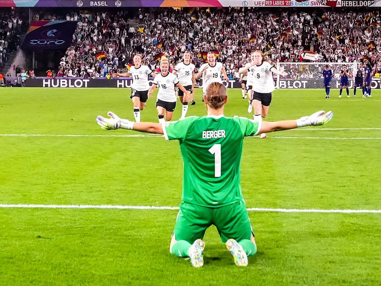 Rückansicht von Ann-Katrin Berger, die vor ihrem Tor kniet und die Arme ihrer Mannschaft entgegenstreckt, die auf sie zurennt.
