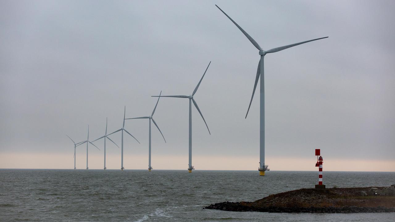 Auf dem Bild sind Offshore-Windkraftwerke in der Nordsee zu sehen. Sie stehen in einer schrägen Linie bis zum Horizont. Im Vordergrund ist eine Landzunge mit einem Leuchtturm.