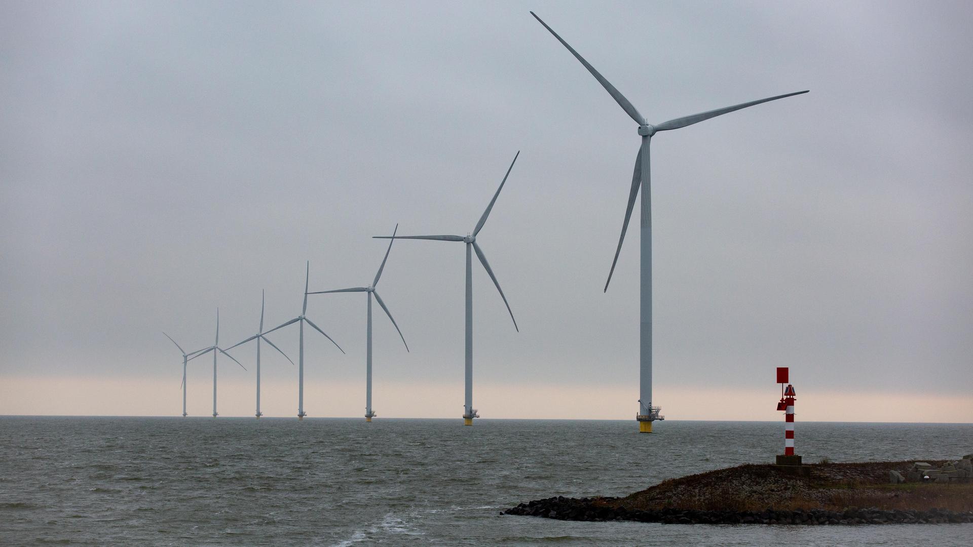 Auf dem Bild sind Offshore-Windkraftwerke in der Nordsee zu sehen. Sie stehen in einer schrägen Linie bis zum Horizont. Im Vordergrund ist eine Landzunge mit einem Leuchtturm.