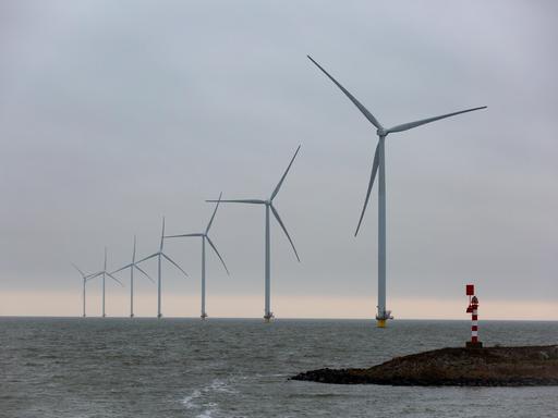 Auf dem Bild sind Offshore-Windkraftwerke in der Nordsee zu sehen. Sie stehen in einer schrägen Linie bis zum Horizont. Im Vordergrund ist eine Landzunge mit einem Leuchtturm.