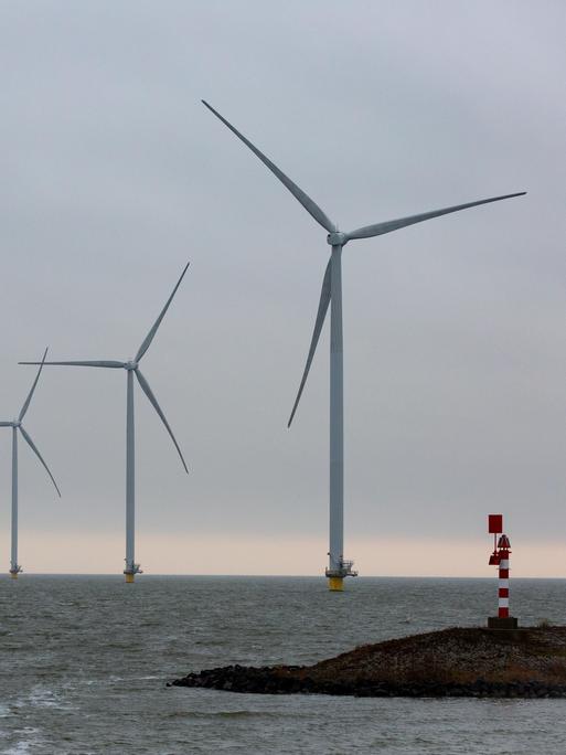 Auf dem Bild sind Offshore-Windkraftwerke in der Nordsee zu sehen. Sie stehen in einer schrägen Linie bis zum Horizont. Im Vordergrund ist eine Landzunge mit einem Leuchtturm.
