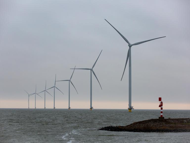 Auf dem Bild sind Offshore-Windkraftwerke in der Nordsee zu sehen. Sie stehen in einer schrägen Linie bis zum Horizont. Im Vordergrund ist eine Landzunge mit einem Leuchtturm.