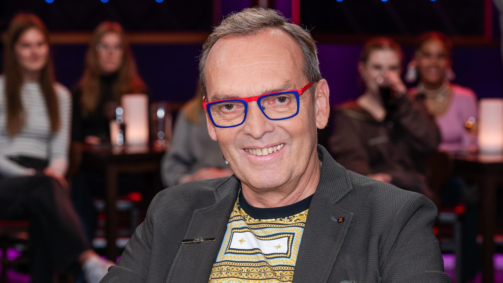 Ein Mann mit dunkelgrauem Haar und markanter, rot-blau gerahmter Brille sitzt lächelnd in einem Fernsehstudio. Er trägt ein graues Sakko und darunter ein gemustertes Shirt. Im Hintergrund verschwommen das Publikum.