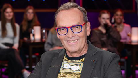 Ein Mann mit dunkelgrauem Haar und markanter, rot-blau gerahmter Brille sitzt lächelnd in einem Fernsehstudio. Er trägt ein graues Sakko und darunter ein gemustertes Shirt. Im Hintergrund verschwommen das Publikum.