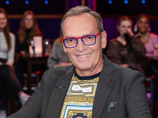 Ein Mann mit dunkelgrauem Haar und markanter, rot-blau gerahmter Brille sitzt lächelnd in einem Fernsehstudio. Er trägt ein graues Sakko und darunter ein gemustertes Shirt. Im Hintergrund verschwommen das Publikum.