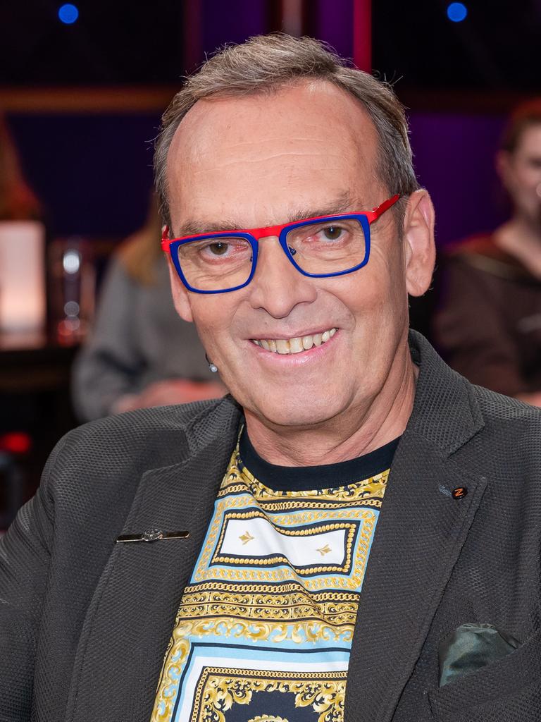 Ein Mann mit dunkelgrauem Haar und markanter, rot-blau gerahmter Brille sitzt lächelnd in einem Fernsehstudio. Er trägt ein graues Sakko und darunter ein gemustertes Shirt. Im Hintergrund verschwommen das Publikum.