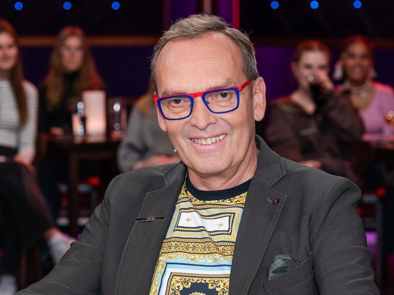 Ein Mann mit dunkelgrauem Haar und markanter, rot-blau gerahmter Brille sitzt lächelnd in einem Fernsehstudio. Er trägt ein graues Sakko und darunter ein gemustertes Shirt. Im Hintergrund verschwommen das Publikum.