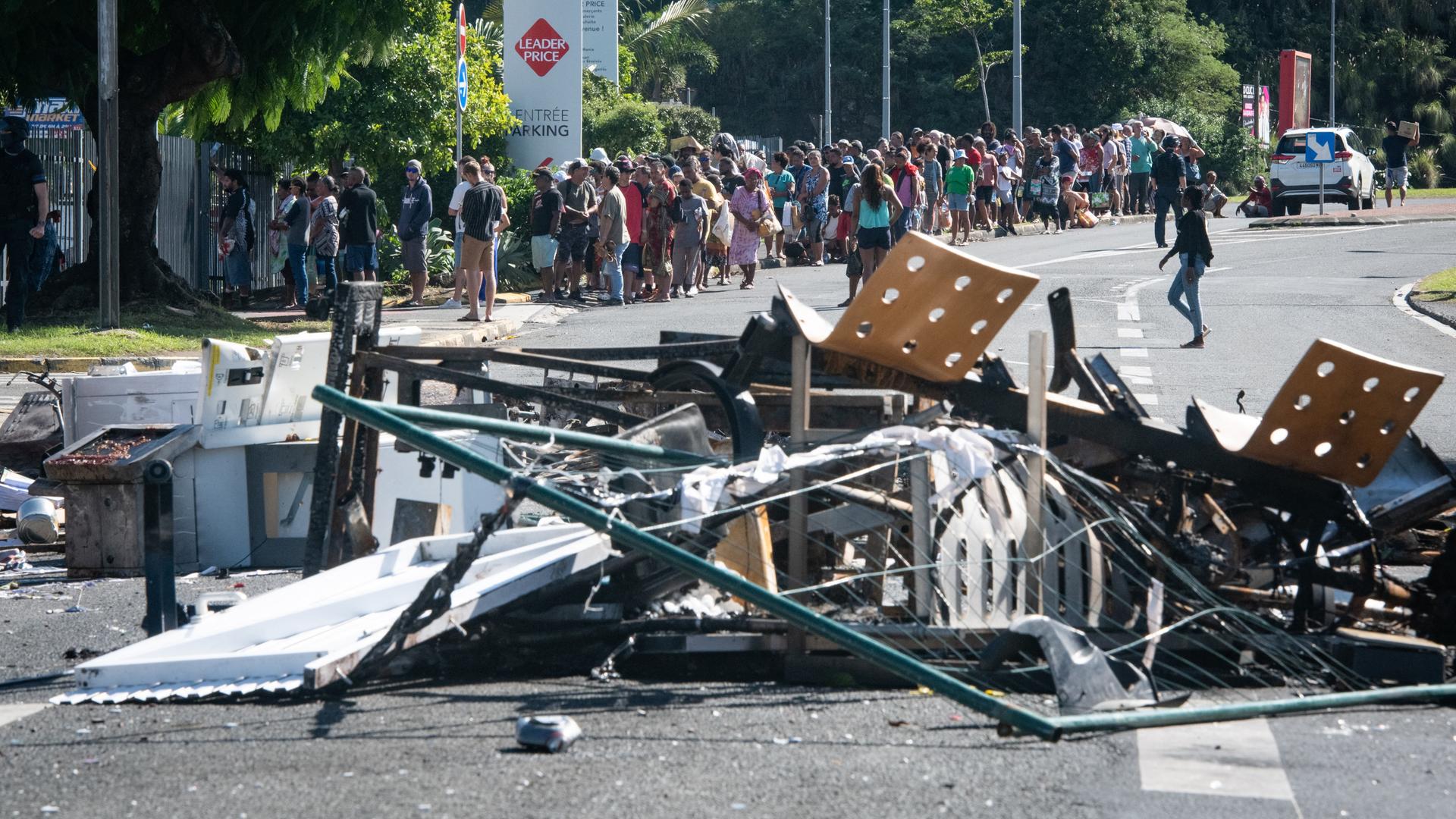 Eine Straßensperre aus Möbeln und anderen Gerätschaften nach nächtlichen Unruhen im Magenta-Bezirk der Hauptstadt Noumea. Im Hintergrund warten Menschen in einer Schlange vor einem Supermarkt.