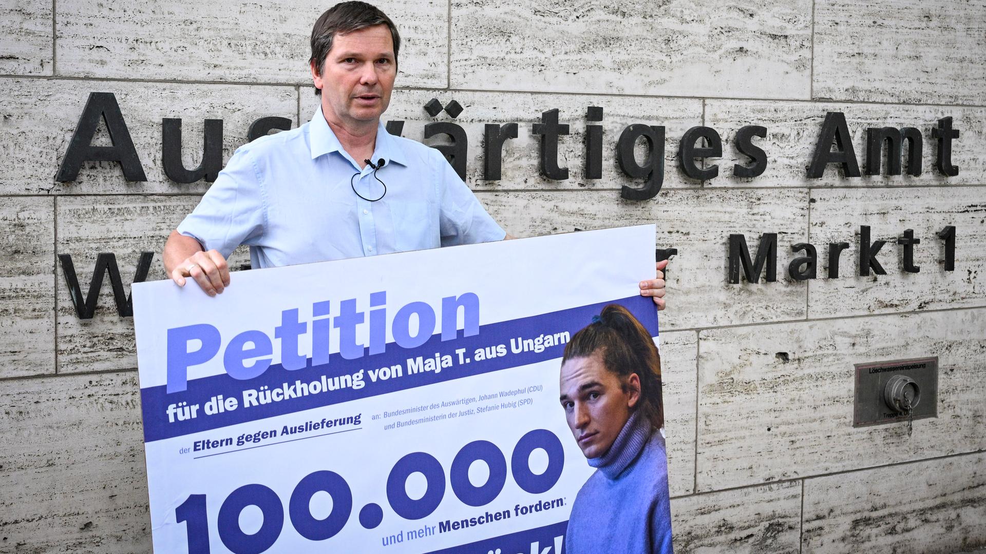 Wolfram Jarosch fordert auf einer Demonstration "Free Maja" die Rückholung seines erwachsenen Kindes Maja T. aus Ungarn. Er steht vor dem Auswärtigen Amt und hält ein Schild hoch. Darauf steht: Für die Rückholung von Maja T aus Ungarn.