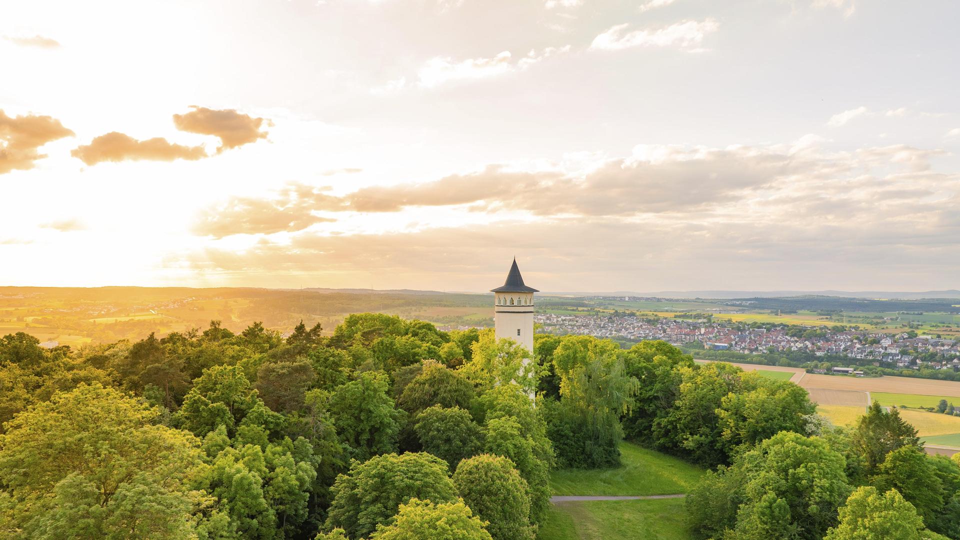 Ein Turm steht umgeben von grünen Hügeln, im Hintergrund ist eine kleine Stadt zu sehen.