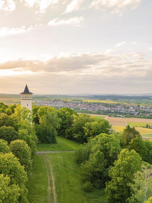 Ein Turm steht umgeben von grünen Hügeln, im Hintergrund ist eine kleine Stadt zu sehen.