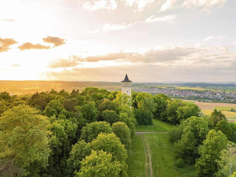 Ein Turm steht umgeben von grünen Hügeln, im Hintergrund ist eine kleine Stadt zu sehen.