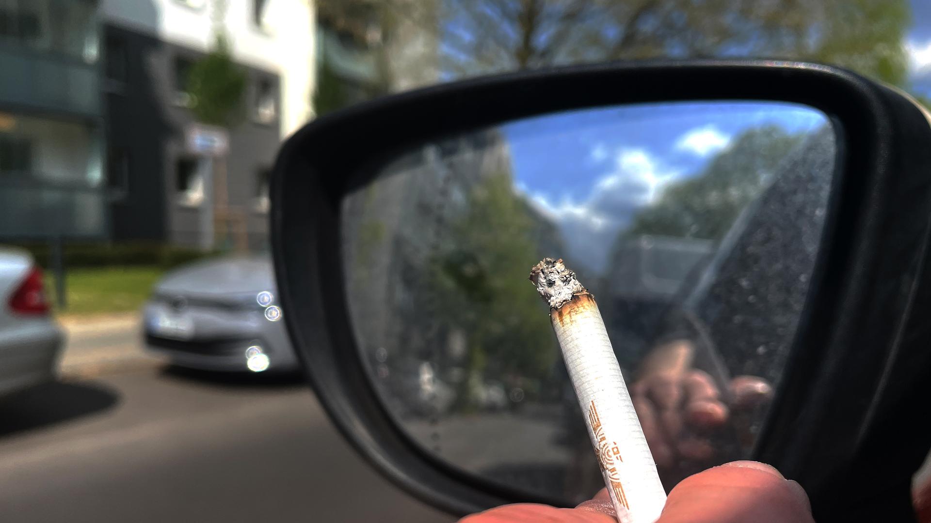 Eine Person hält eine Zigarette beim Autofahren aus dem Fenster.
Eine Person hält eine Zigarette beim Autofahren aus dem Fenster.