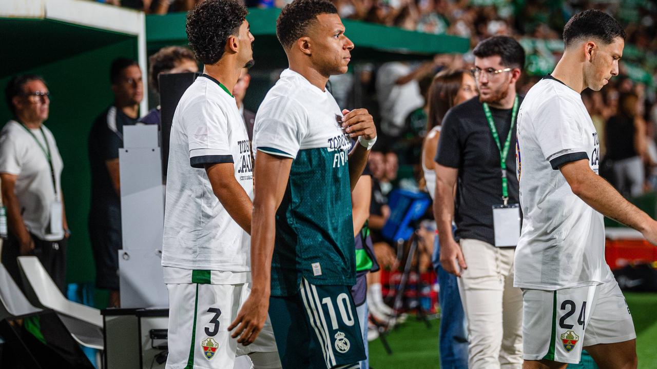 Mehrere Fußball-Spieler laufen in ein Stadion ein. Einer von ihnen hat ein T-Shirt zwar übergestreift, aber nicht komplett heruntergezogen.