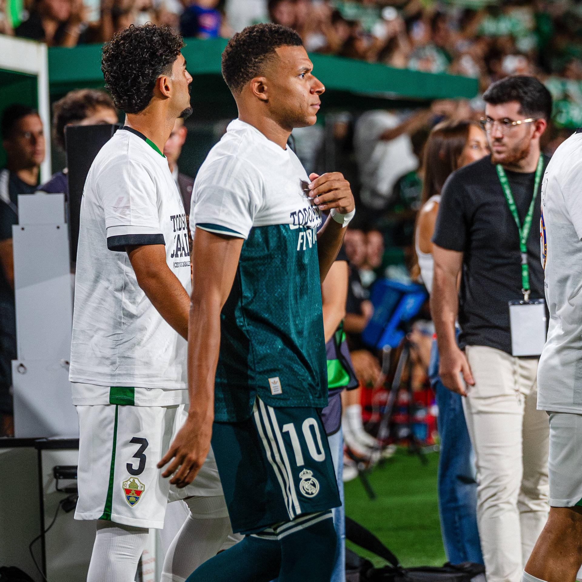 Mehrere Fußball-Spieler laufen in ein Stadion ein. Einer von ihnen hat ein T-Shirt zwar übergestreift, aber nicht komplett heruntergezogen.