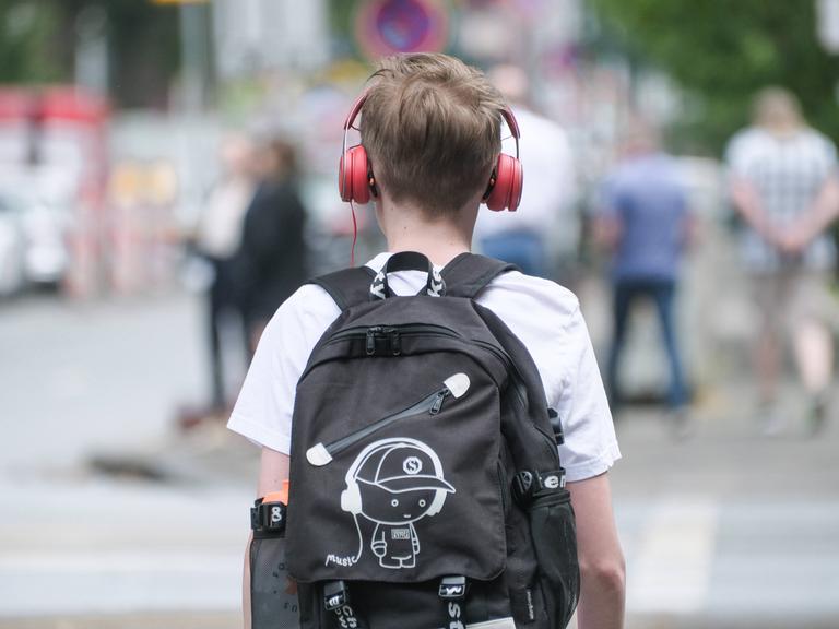 Ein Junge ist von hinten zu sehen. Er trägt Kopfhörer und einen schwarzen Rucksack, auf dem ebenfalls ein Junge mit Kopfhörer zu sehen ist. 