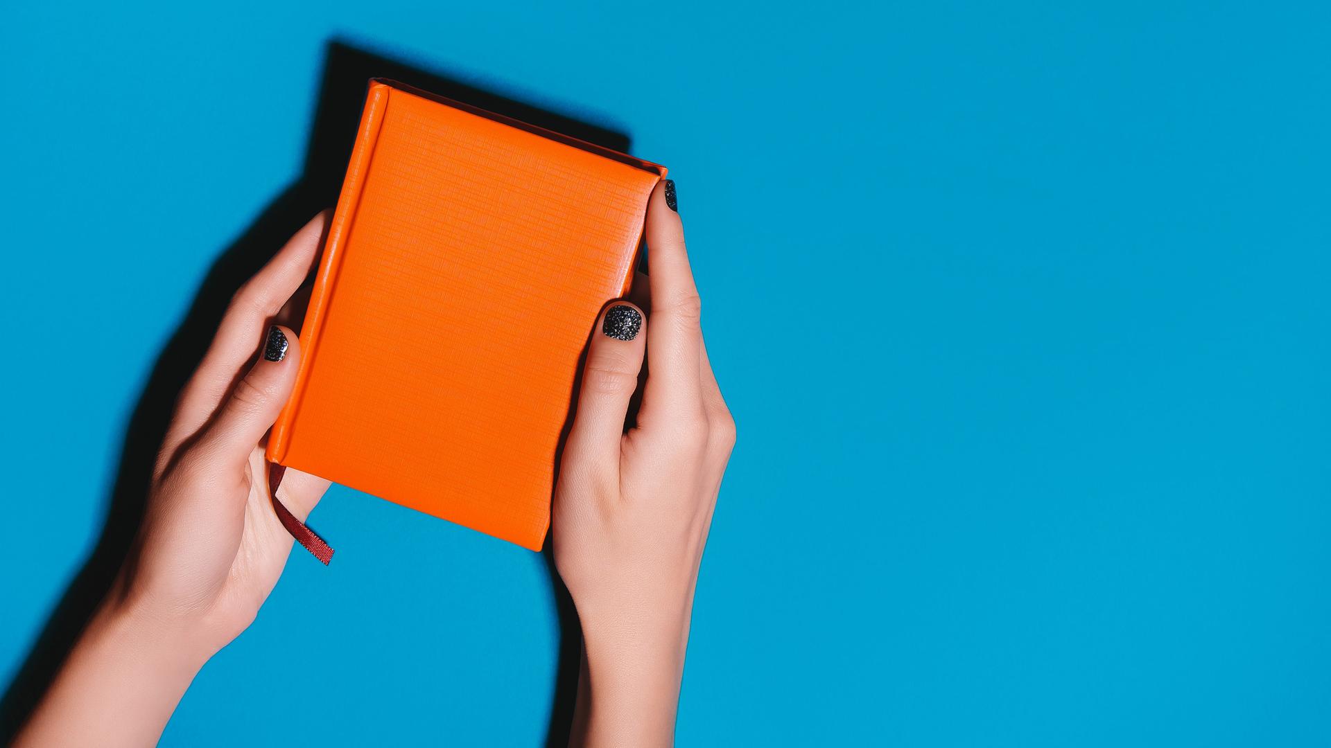 Eine Person mit dunkel lackierten Fingernägeln hält ein orangenes Tagebuch über blauem Hintergrund. Eine Person mit dunkel lackierten Fingernägeln hält ein orangenes Tagebuch über blauem Hintergrund.
