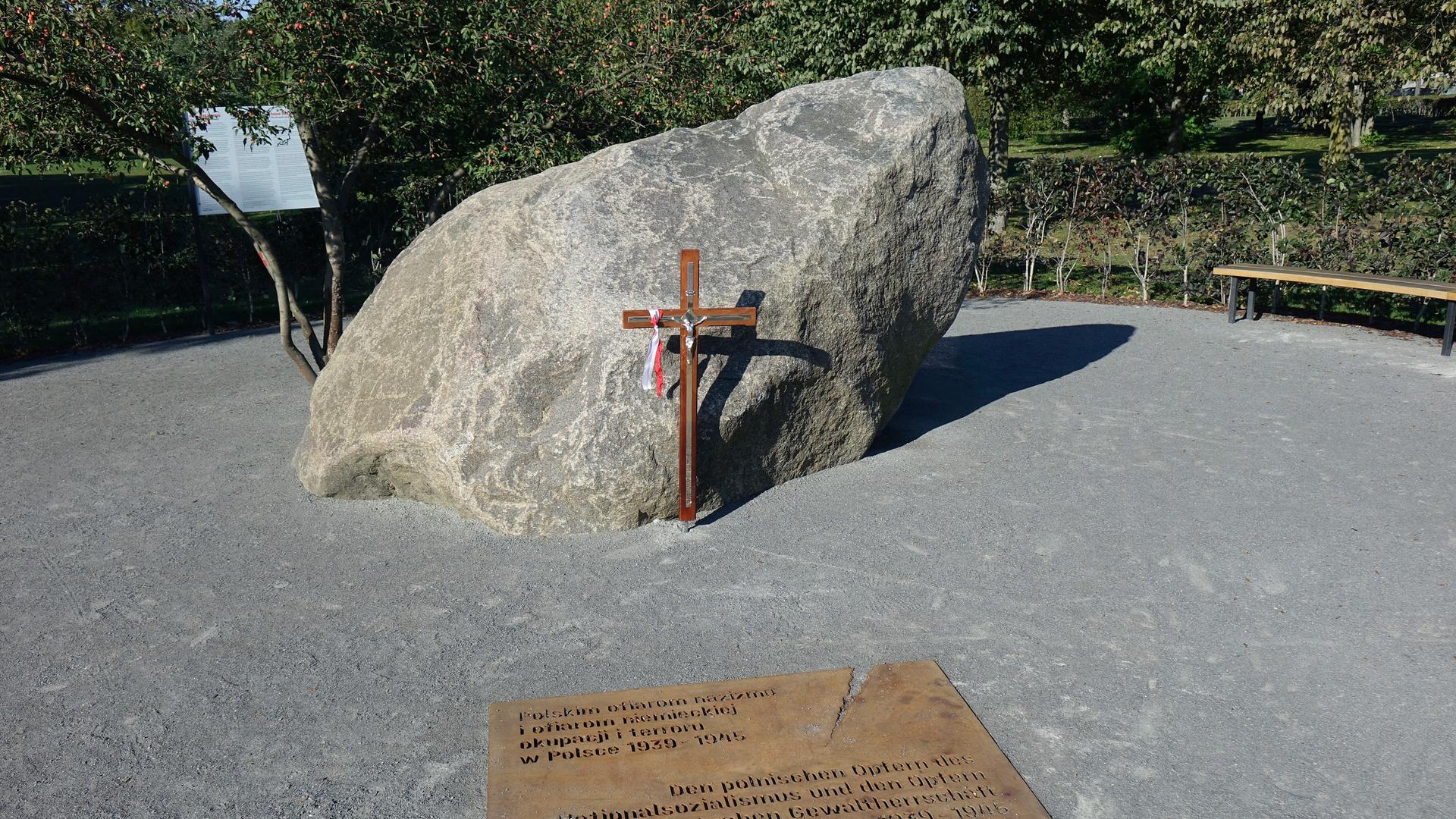 Ein Kreuz steht vor einem großen Stein. Davor ist eine Tafel im Boden eingelassen mit einer Inschrift.