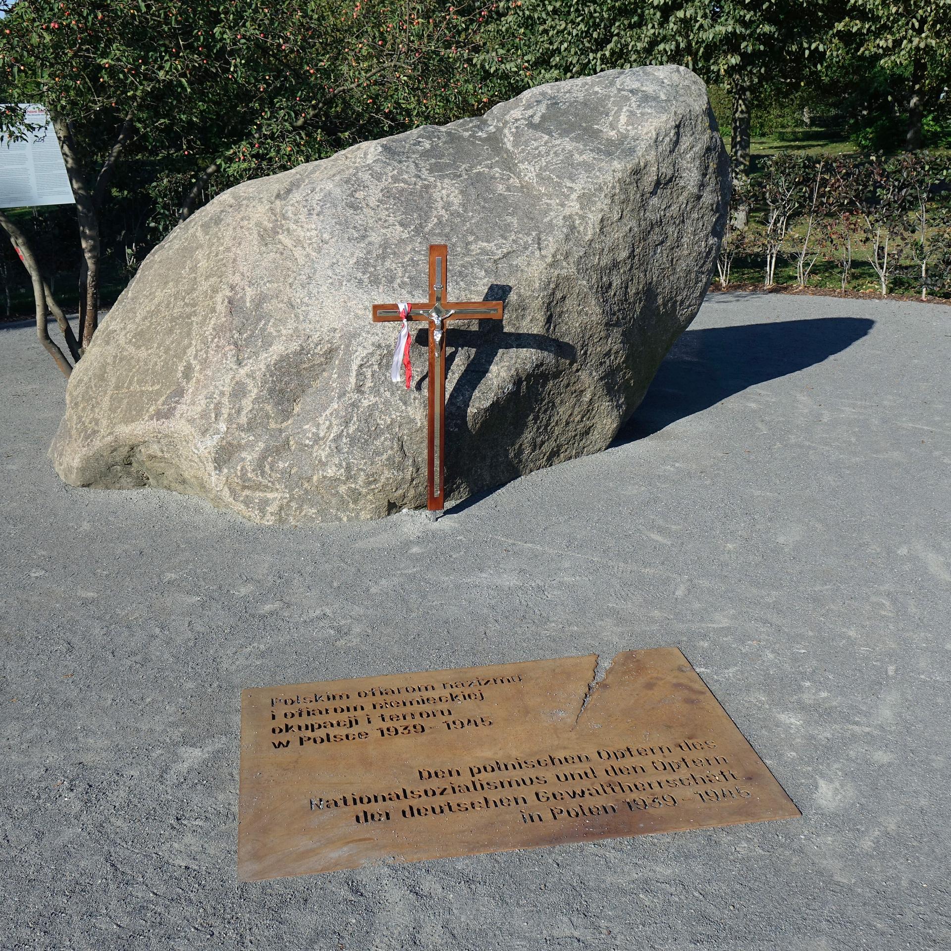 Ein Kreuz steht vor einem großen Stein. Davor ist eine Tafel im Boden eingelassen mit einer Inschrift.