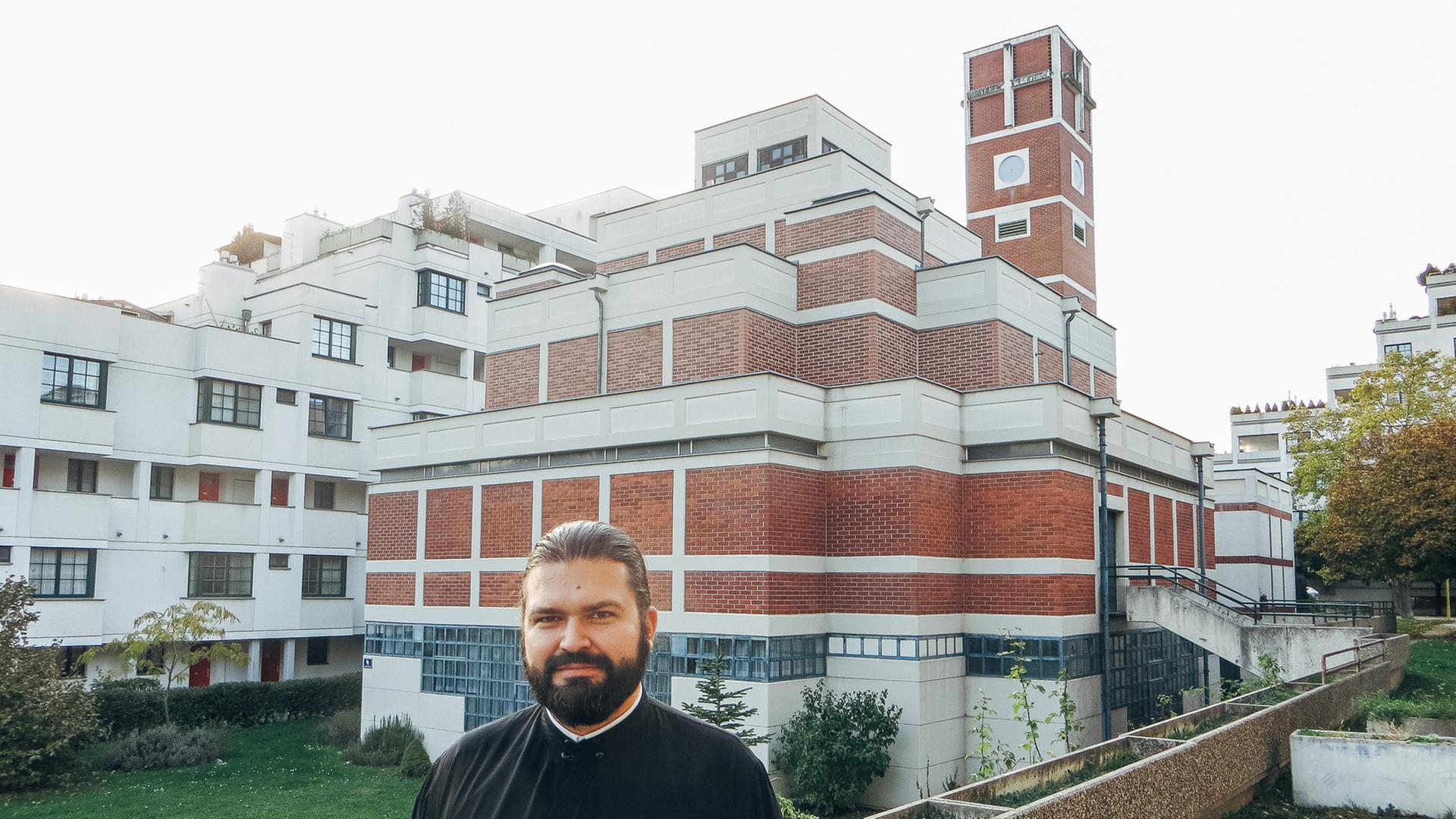 Priester Filip Milunovic vor seiner Kirche Am Schöpfwerk in Wien. Er trägt Talar, hat einen Vollbart und die dunklen Haare nach hinten gekämmt. Die Kirche setzt sich aus kubischen Elementen zusammen, zum Teil sind die Wände verputzt, zum Teil sieht man Sichtziegelmauerwerk.