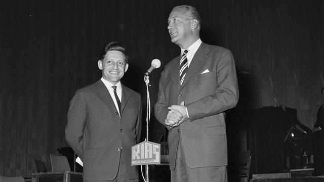 Schwarz-Weiß-Aufnahme von zwei Männern im Anzug auf einer Bühne hinter einem RIAS-Mikrofon