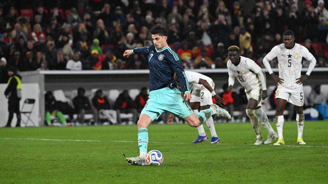 Ein Mann in blauem Trikot schießt auf einen Fußball, im Hintergrund stehen mehrere Spieler in weißen Trikots.