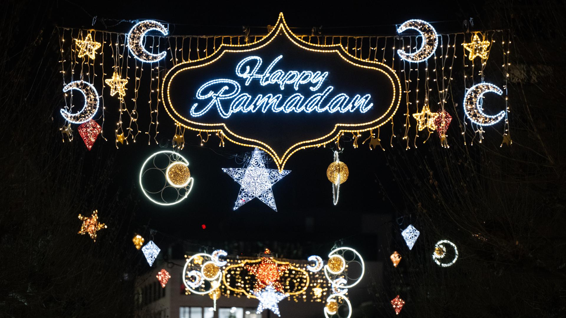 Zu sehen ist der blau-silber leuchtende Schriftzug „Happy Ramadan“ in einer gold leuchtenden geschwungen Umrandung. Daneben leuchten Halbmonde und Sterne. Im Hintergrund ist der dunkle Nachthimmel zu sehen. 
