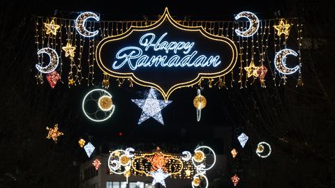 Zu sehen ist der blau-silber leuchtende Schriftzug „Happy Ramadan“ in einer gold leuchtenden geschwungen Umrandung. Daneben leuchten Halbmonde und Sterne. Im Hintergrund ist der dunkle Nachthimmel zu sehen. 
