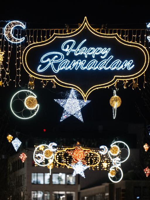 Zu sehen ist der blau-silber leuchtende Schriftzug „Happy Ramadan“ in einer gold leuchtenden geschwungen Umrandung. Daneben leuchten Halbmonde und Sterne. Im Hintergrund ist der dunkle Nachthimmel zu sehen.
Zu sehen ist der blau-silber leuchtende Schriftzug „Happy Ramadan“ in einer gold leuchtenden geschwungen Umrandung. Daneben leuchten Halbmonde und Sterne. Im Hintergrund ist der dunkle Nachthimmel zu sehen.
