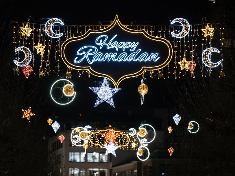 Zu sehen ist der blau-silber leuchtende Schriftzug „Happy Ramadan“ in einer gold leuchtenden geschwungen Umrandung. Daneben leuchten Halbmonde und Sterne. Im Hintergrund ist der dunkle Nachthimmel zu sehen.
Zu sehen ist der blau-silber leuchtende Schriftzug „Happy Ramadan“ in einer gold leuchtenden geschwungen Umrandung. Daneben leuchten Halbmonde und Sterne. Im Hintergrund ist der dunkle Nachthimmel zu sehen.