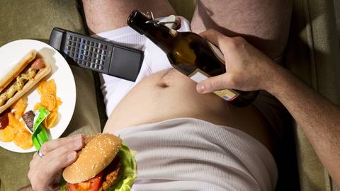 Ein Mann sitzt in der Unterhose auf der Couch mit einer Bierflasche und einem Hamburger in seinen Händen.