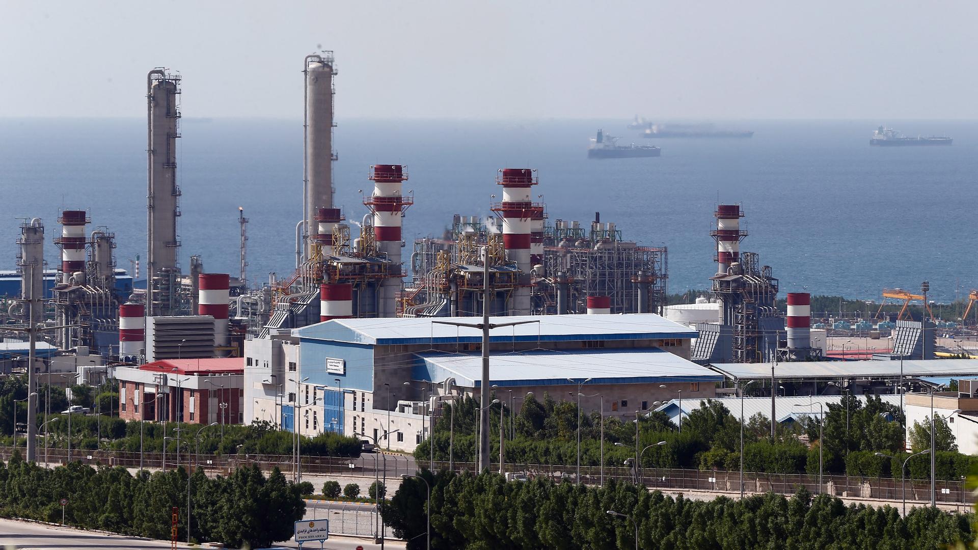 Das Bild zeigt eine Industrieanlage mit großen Rohren und anderen Konstruktionen aus Metall und Beton. Im Hintergrund ist ein Meer zu erkennen.