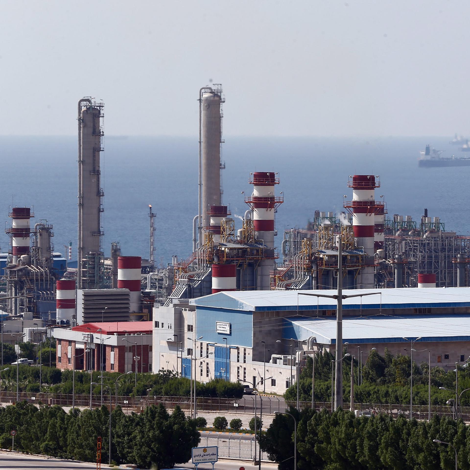 Das Bild zeigt eine Industrieanlage mit großen Rohren und anderen Konstruktionen aus Metall und Beton. Im Hintergrund ist ein Meer zu erkennen.