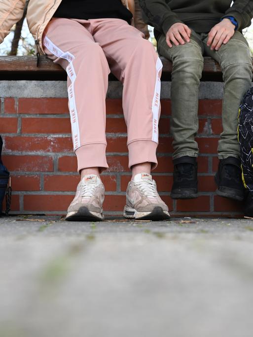 Eine Schülerin sitzt in einer Jogginghose auf dem Schulhof einer Schule. Abgebildet sind nur die Beine. 