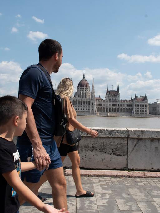 Eine Familie läuft am Ufer der Donau in Budapest entlang, auf der anderen Flussseite ist das Parlamentsgebäude zu sehen