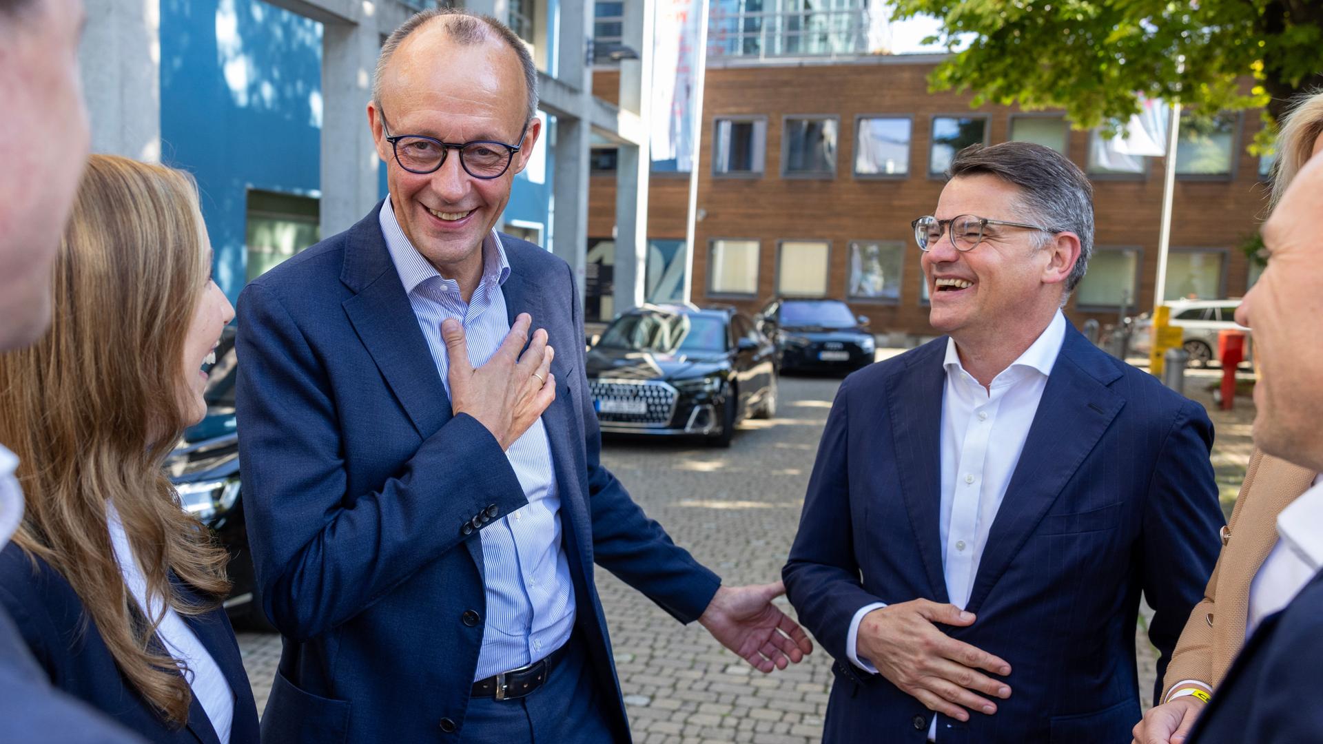 Merz steht grinsend auf einer gepflasterten Straße vor Gebäuden und sagt etwas; Rhein steht neben ihm und lacht. Daneben weitere Personen.