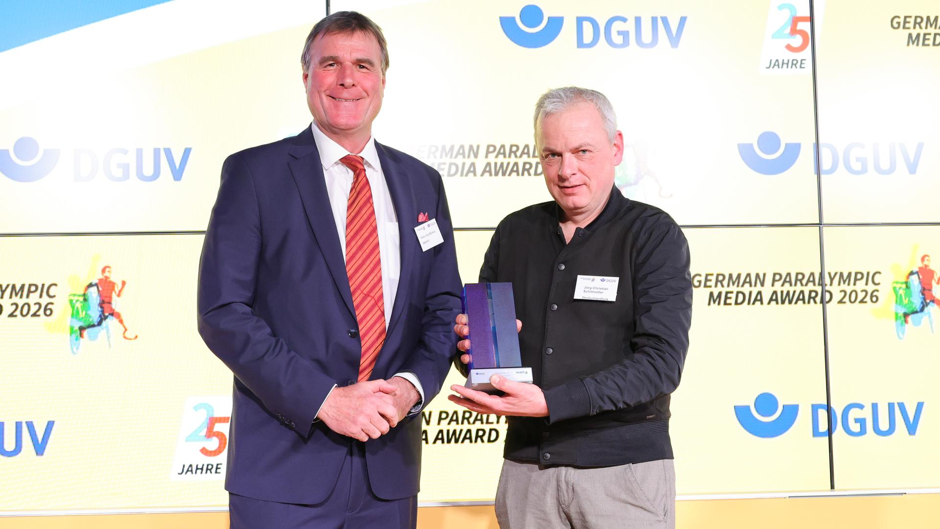 Jörg-Christian Schillmöller (rechts) und der Präsident des Deutschen Behinderten-Sport-Verbands, Hans-Jörg Michels, stehen auf einer Bühne in Berlin. Jörg-Christian Schillmöller hält den Preis in der Hand.