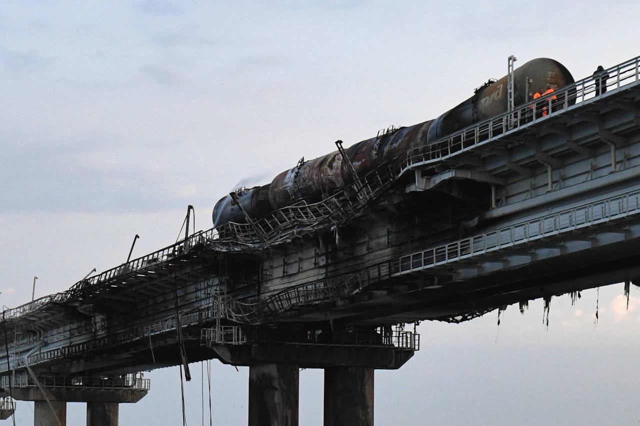 Blick von unten auf mehrere ausgebrannte Güterwaggons auf einem beschädigten Brückenabschnitt unter grauem Himmel. 