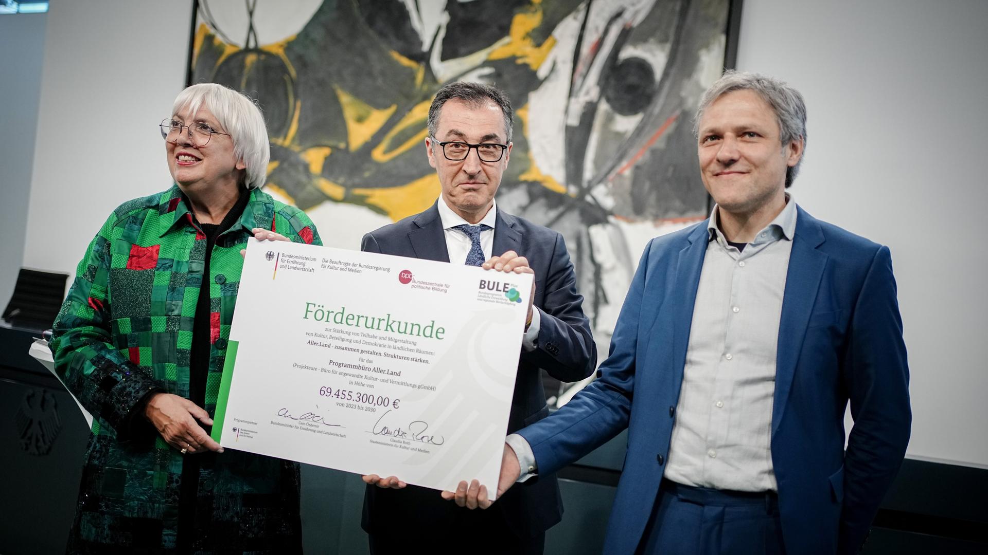 Kulturstaatsministerin Roth und Bundeslandwirtschaftsminister Özdemir übergeben einen großformatigen Förderbescheid an Samo Darian, Leiter des Programmbüros Aller.Land.