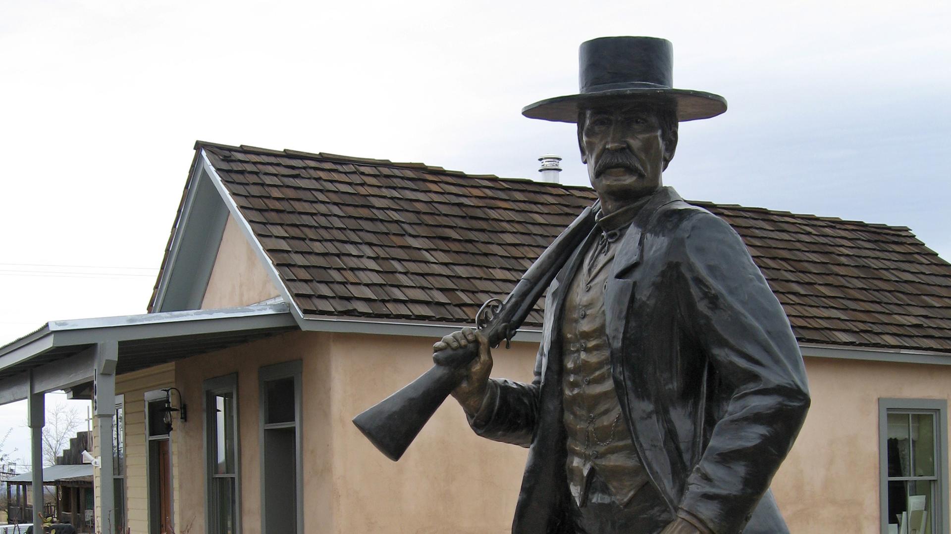 Eine Bronze-Statue des Künstlers Tim Trask erinnert einem Friedhof in Arizona an den Revolverhelden Wyatt Earp. Eine Lebensgroße Figur mit Mantel steht auf einem Sockel und hält ein Gewehr. 