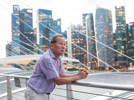 Singapur – Asiatisches Porträt: Ein älterer Mann blickt auf die modern Hochhauslandschaft in Singapur und denkt über die Zukunft nach