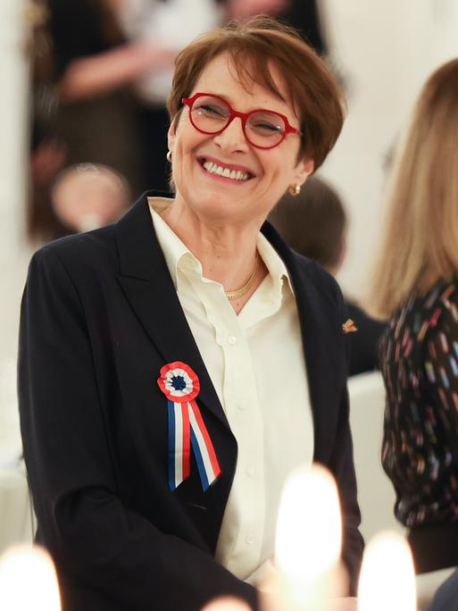 Hélène Miard-Delacroix trägt eine rote Brille und eine Brosche in den französischen Nationalfarben. Sie schaut zur Seite und lacht die Person links im Bild (Bundestagspräsidentin Bärbel Bas, nur von hinten zu sehen) an. 