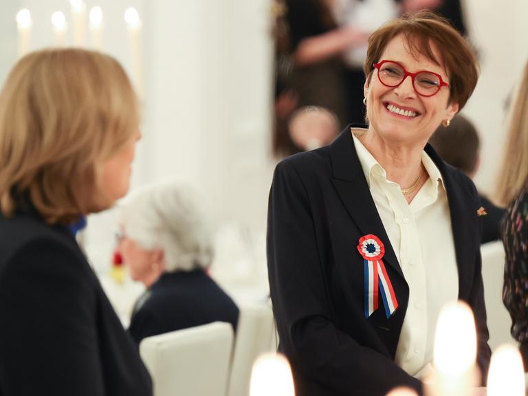 Hélène Miard-Delacroix trägt eine rote Brille und eine Brosche in den französischen Nationalfarben. Sie schaut zur Seite und lacht die Person links im Bild (Bundestagspräsidentin Bärbel Bas, nur von hinten zu sehen) an. 