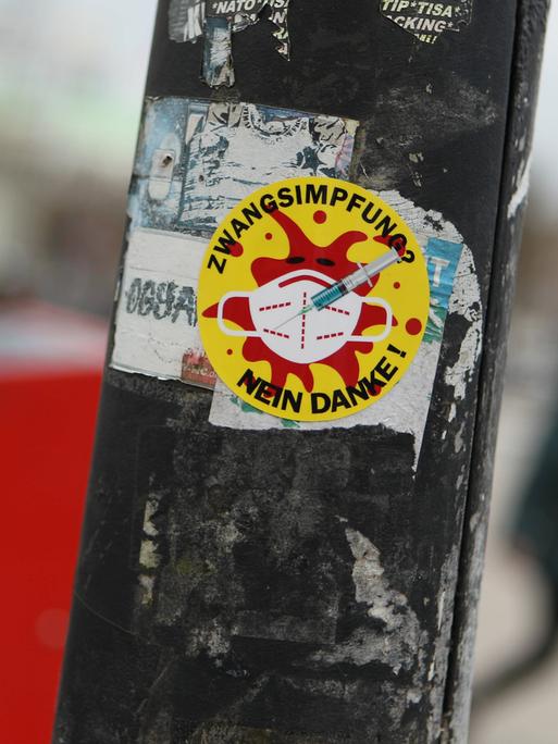 Sticker an einem Laternenpfahl, auf dem steht "Zwangs-Impfung? Nein danke". Das Motiv, ein rotes Coronavirus auf gelben Grund, erinnert an die Anti-Atomkraft-Aufkleber mit dem Text "Atomkraft? Nein danke".