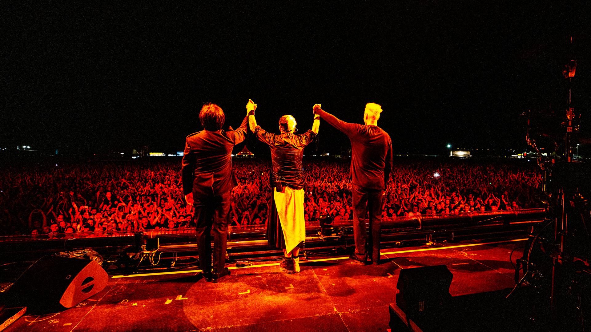 Drei Mitglieder der Band "Die Ärzte" sind von hinten zu sehen. Sie stehen auf einer Bühne und halten sich an den Händen, um sich vor einem großen Publikum zu verbeugen.