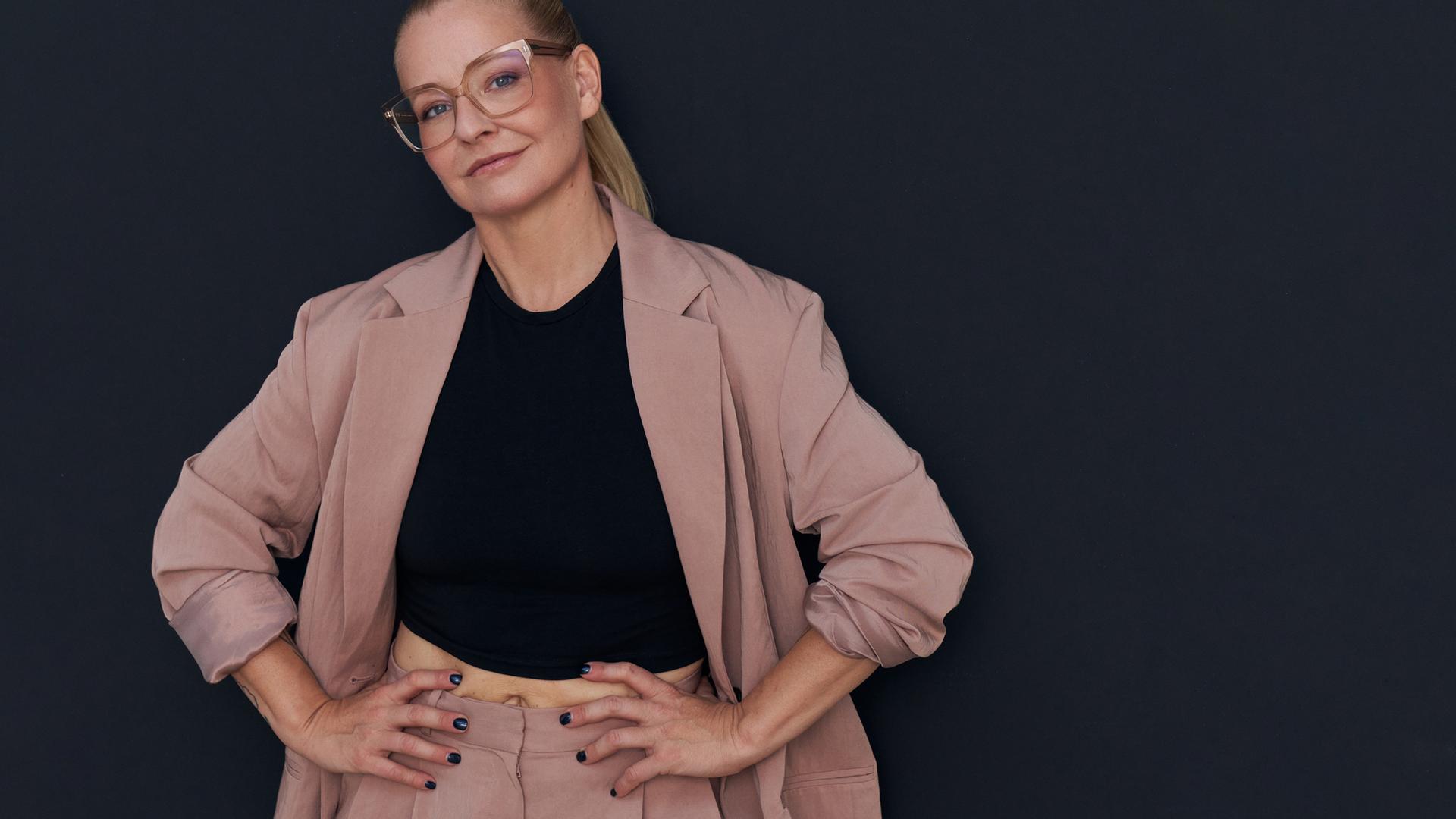 Die Autorin Franziska Hohmann mit Brille trägt einen rosafarbenden Anzug, schaut in die Kamera und hat ihre Hände in ihre Hüften gestützt