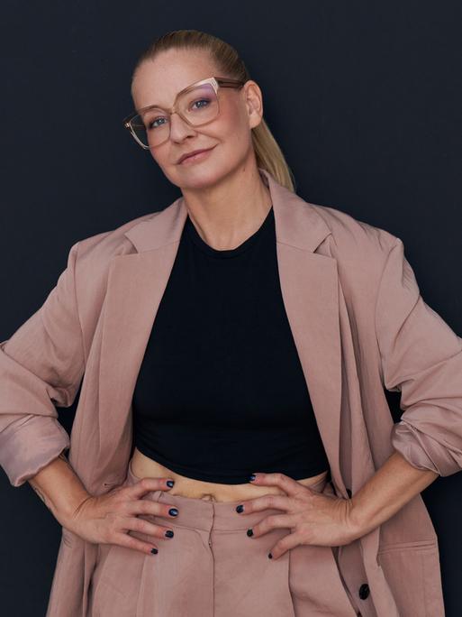 Die Autorin Franziska Hohmann mit Brille trägt einen rosafarbenden Anzug, schaut in die Kamera und hat ihre Hände in ihre Hüften gestützt