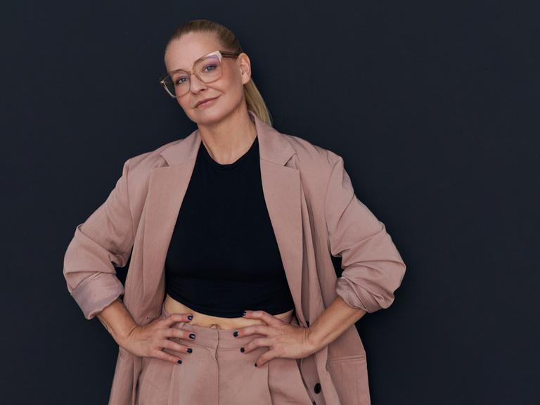 Die Autorin Franziska Hohmann mit Brille trägt einen rosafarbenden Anzug, schaut in die Kamera und hat ihre Hände in ihre Hüften gestützt