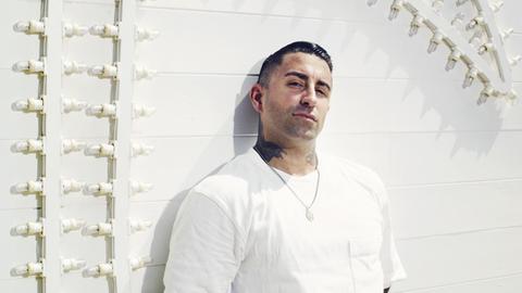 Sammy Amara, Sänger der Band Broilers, lehnt an einer weißen Wand und blickt herausfordernd in die Kamera.