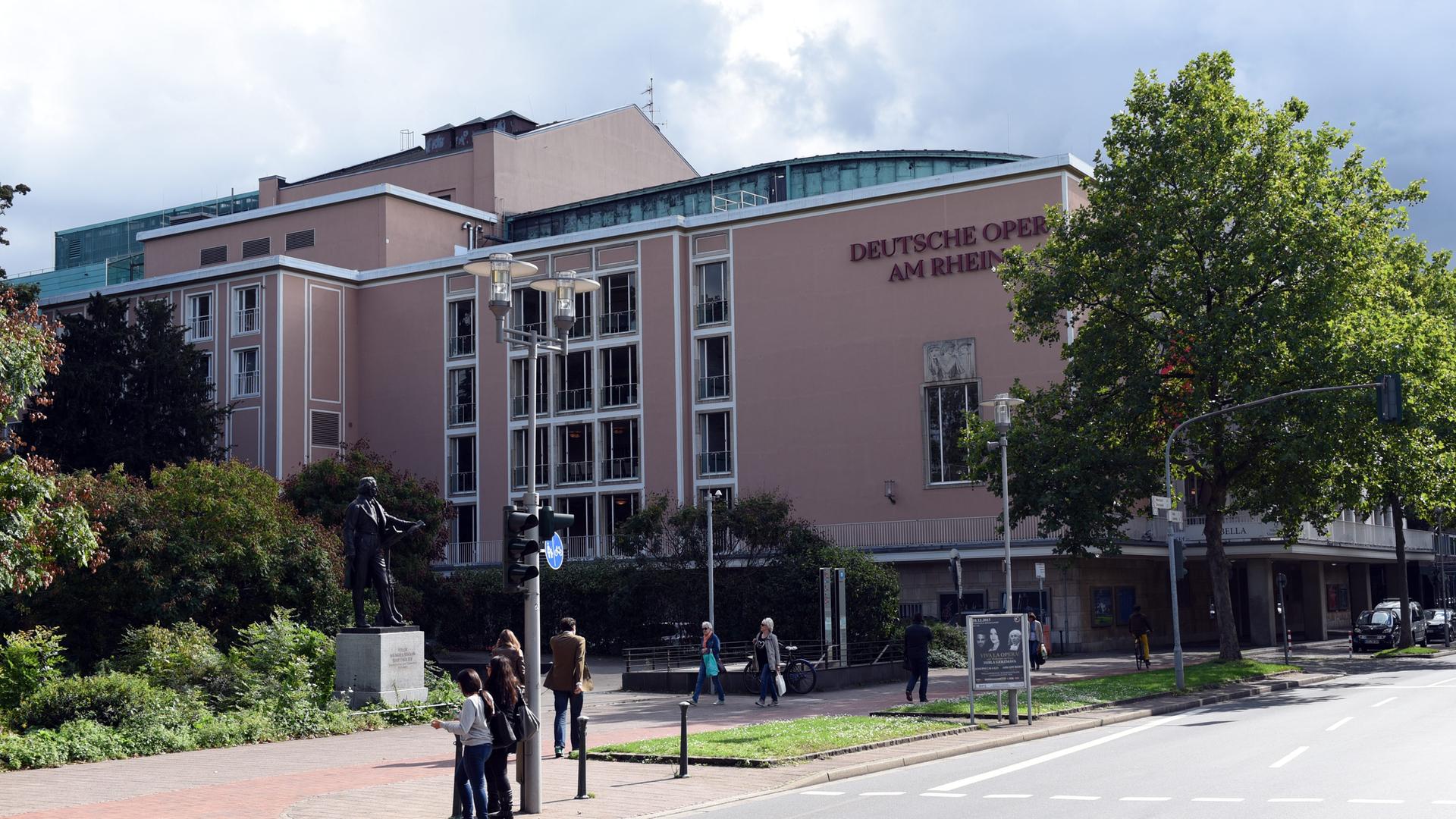 Das Opernhaus in Düsseldorf- ein großes ockerfarbenes Gebäude. Daneben ist eine Straße. Im Vordergund sind Passanten zu sehen. 